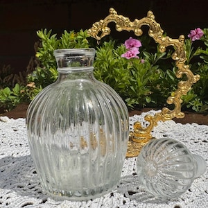 Puede incluir: Un frasco de perfume de vidrio transparente con acanaladuras verticales y un tapón de vidrio redondo a juego. El frasco está colocado junto a un soporte de metal dorado curvo y ornamentado con volutas y detalles decorativos.