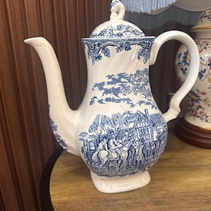 Może przedstawiać: Biały ceramiczny imbryk z niebieskim wzorem przedstawiającym wiejską scenę z postaciami na koniach, drzewami i budynkiem. Imbryk posiada zakrzywiony uchwyt, długą wylewkę oraz pasującą pokrywkę z ozdobnym uchwytem w kształcie gałki.