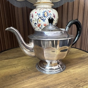 Op de afbeelding: Een gepolijste theepot van zilverkleurig metaal met een gefacetteerd, veelzijdig lichaam, een gebogen tuit en een zwart handvat. Het deksel is voorzien van een kleine zwarte knop. De theepot rust op een ronde voet en reflecteert het licht.