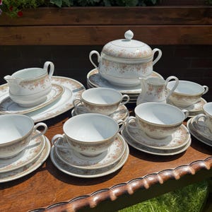 May include: A white porcelain tea set featuring a delicate floral pattern in shades of pink and green with gold trim. The set includes a lidded tureen, a creamer, multiple teacups with matching saucers, and serving plates arranged on a wooden table.