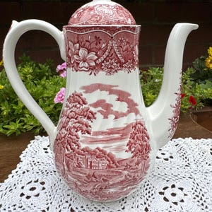 Può includere: Una teiera in ceramica bianca con un motivo a trasferimento rosso. Il corpo presenta una scena paesaggistica dettagliata con alberi, un edificio e un ponte. Il coperchio e il bordo superiore sono decorati con motivi floreali. È dotata di un manico e un beccuccio curvi.