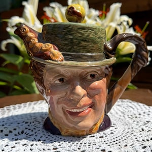 May include: A ceramic character teapot shaped like a person wearing a green textured hat. The spout features a brown decorative element, and the handle is shaped like a twisted branch. The lid has a small round knob with a brown detail.