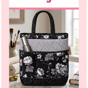May include: A quilted cloth tote bag featuring a black and white floral pattern on the main body, a gray quilted upper section, and black leopard print trim on the handles and base. The bag includes exterior pockets and a silver metallic object.