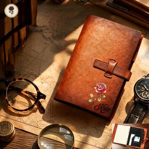 May include: A brown leather journal with a strap closure, featuring a printed red rose illustration and the name Kate in cursive script. The journal is displayed alongside tortoiseshell glasses, a wristwatch, and a magnifying glass.