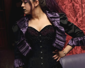 May include: A woman wearing a black corset with purple trim and a black jacket with purple ruffles. The corset has a lace-up front and the jacket has long sleeves. The woman is standing in front of a red patterned background.