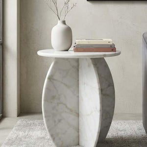May include: A white marble side table with a circular top and a base composed of two intersecting vertical panels. A matte white ceramic vase containing dried stems and a stack of three books are placed on the surface of the table.
