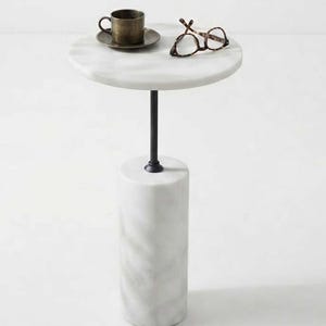 May include: A round white marble side table with a central black metal post connecting a cylindrical marble base to a circular marble top. On the table sits a metallic brown cup on a matching saucer and a pair of brown tortoiseshell spectacles.