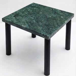 Pode incluir: Uma mesa lateral quadrada com um tampo de mármore verde polido com veios e textura naturais. A mesa é suportada por quatro pernas cilíndricas de metal preto mate posicionadas em cada canto, proporcionando uma base estável.