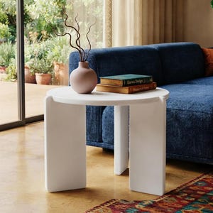 May include: A white round side table with three wide, flat legs. On the table sits a matte pink vase containing thin, dark, twisted branches and two stacked books with the visible text Eames Design and Modernist Forms on their spines.