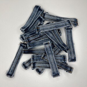 Può includere: Una collezione di piccoli passanti per cintura in denim blu di forma rettangolare, caratterizzati da una classica trama a saia e cuciture marroni visibili lungo i bordi. Queste strisce di tessuto sono disposte in un mucchio sparso.