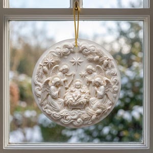 May include: A circular off-white ornament featuring a relief carving of a nativity scene with a central infant, surrounding angels, and a star. The ornament is suspended by a thin gold cord and displays intricate details of clouds and pine branches.