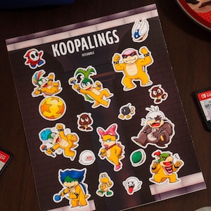 Può includere: Un foglio di adesivi rettangolare intitolato KOOPALINGS KITSABBLE con vari adesivi colorati di personaggi dei cartoni animati, tra cui creature gialle, blu e marroni, una figura con maschera rossa, una palla gialla, un guscio verde e un fantasma bianco.
