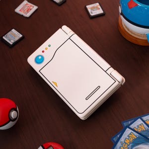 May include: A white handheld gaming console featuring a stylized design resembling a Pok&eacute; Ball, with a blue circular button, three small indicator lights, and a yellow triangle accent on the top cover, surrounded by various small game cartridges and cards.