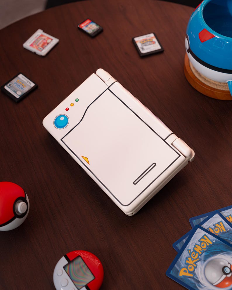 May include: A white handheld gaming console featuring a stylized design resembling a Pok&eacute; Ball, with a blue circular button, three small indicator lights, and a yellow triangle accent on the top cover, surrounded by various small game cartridges and cards.