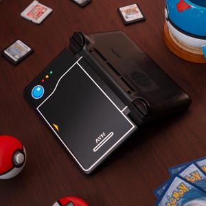 May include: A black handheld gaming console with a Pok&eacute; Ball design on the lid, featuring the text AYN. The console is surrounded by three small game cartridges, a blue and white Pok&eacute; Ball mug, a red and white Pok&eacute; Ball, a digital pet, and cards.