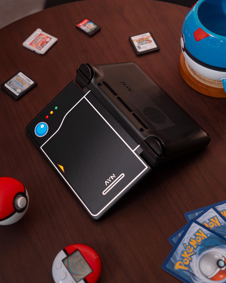May include: A black handheld gaming console with a Pok&eacute; Ball design on the lid, featuring the text AYN. The console is surrounded by three small game cartridges, a blue and white Pok&eacute; Ball mug, a red and white Pok&eacute; Ball, a digital pet, and cards.
