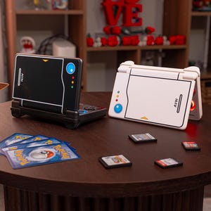 May include: Two handheld gaming consoles, one black and one white, featuring a stylized design with a blue circle and three small colored dots. Beside them are a small brown Eevee figure, several trading cards, four game cartridges, and a red-and-white ball.
