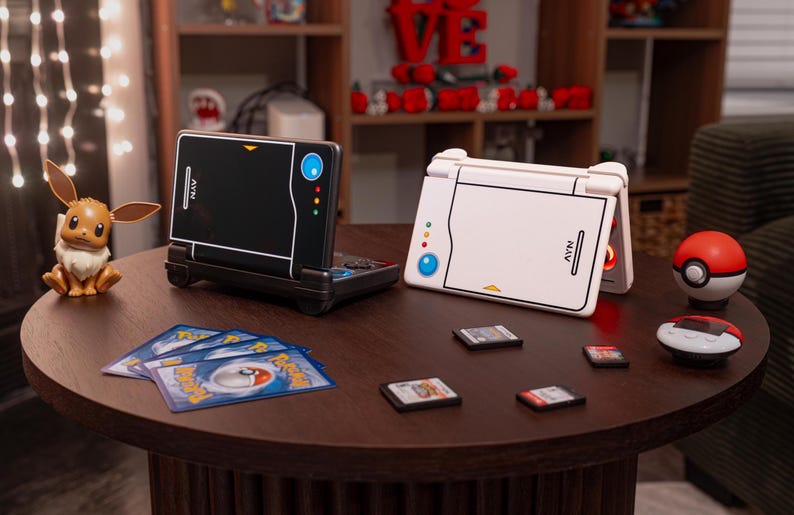May include: Two handheld gaming consoles, one black and one white, featuring a stylized design with a blue circle and three small colored dots. Beside them are a small brown Eevee figure, several trading cards, four game cartridges, and a red-and-white ball.