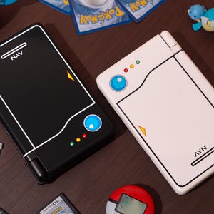 May include: Two handheld gaming consoles, one black and one white, featuring a stylized design with a blue circular button, three small colored lights, and the text AYN printed on the surface. The consoles are surrounded by various small gaming accessories.