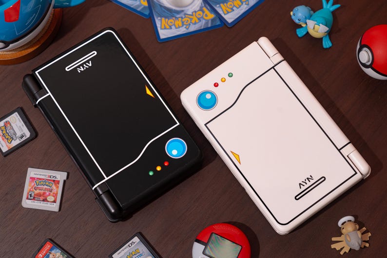 May include: Two handheld gaming consoles, one black and one white, featuring a stylized design with a blue circular button, three small colored lights, and the text AYN printed on the surface. The consoles are surrounded by various small gaming accessories.