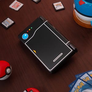 May include: A black handheld gaming console featuring a white outline design resembling a Pok&eacute; Ball, with a blue circular button, three small colored indicator lights, and a small orange triangle accent on the front cover of the closed device.