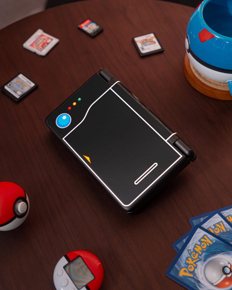 May include: A black handheld gaming console featuring a white outline design resembling a Pok&eacute; Ball, with a blue circular button, three small colored indicator lights, and a small orange triangle accent on the front cover of the closed device.