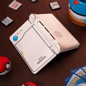 May include: A white handheld gaming console with a Pokedex-inspired design, featuring a blue circular button, three small indicator lights, and the text AYN printed on the front. The device is angled open, showing a speaker grille and ventilation slots.