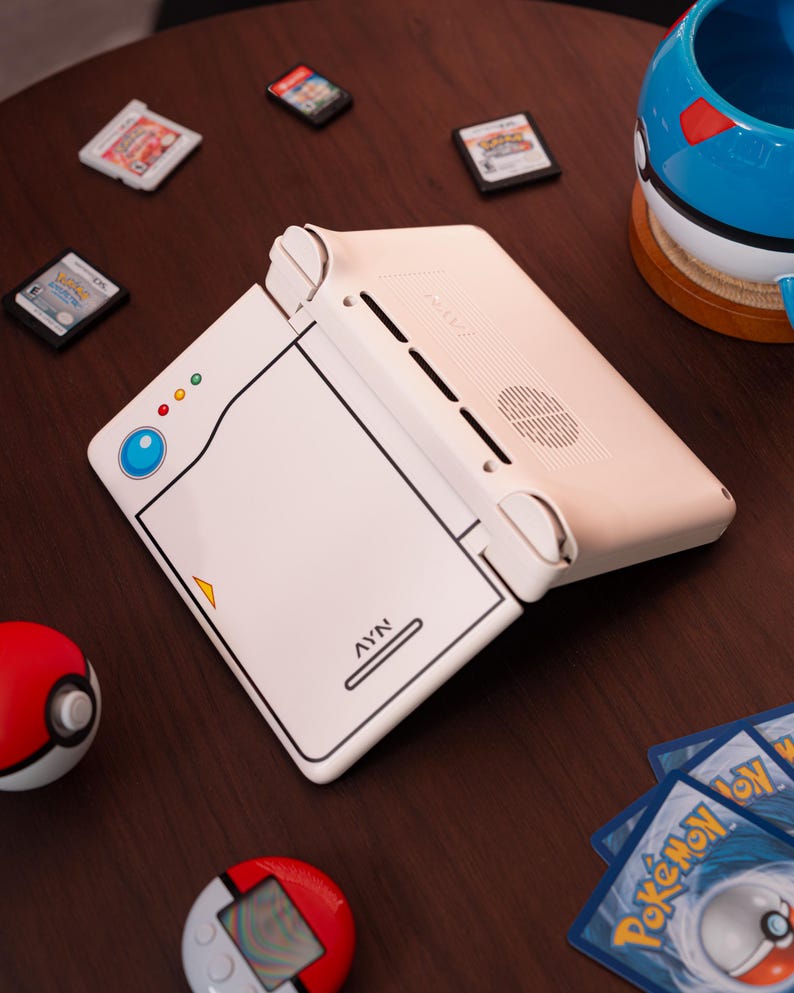 May include: A white handheld gaming console with a Pokedex-inspired design, featuring a blue circular button, three small indicator lights, and the text AYN printed on the front. The device is angled open, showing a speaker grille and ventilation slots.