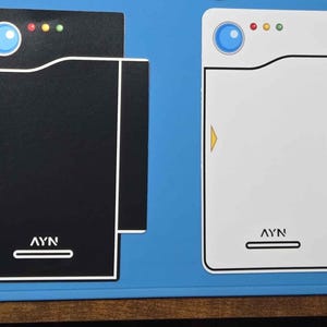May include: Two rectangular flat stickers featuring a stylized design with a blue circle, three small colored dots, a yellow arrow, and the text AYN at the bottom. One sticker is black with a white outline, and the other is white with a black outline.