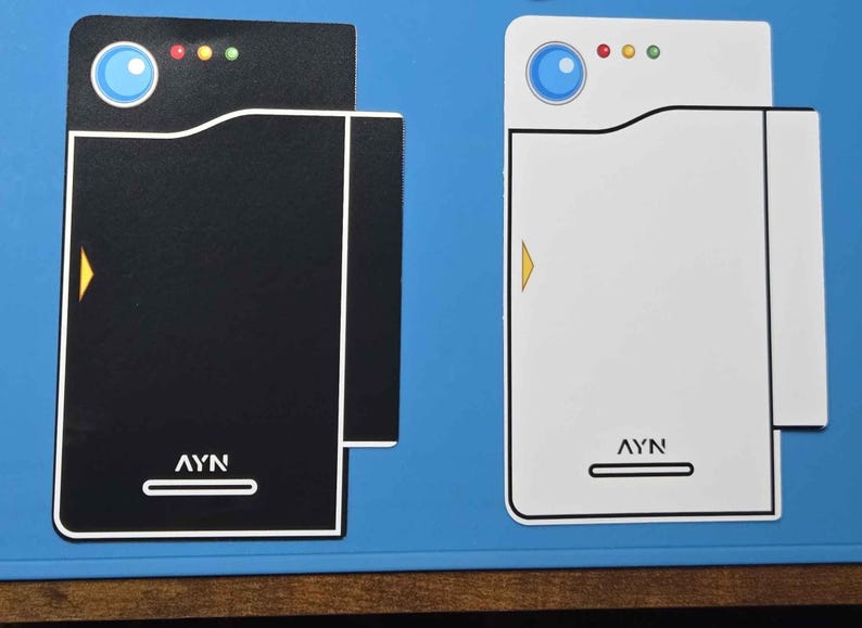 May include: Two rectangular flat stickers featuring a stylized design with a blue circle, three small colored dots, a yellow arrow, and the text AYN at the bottom. One sticker is black with a white outline, and the other is white with a black outline.