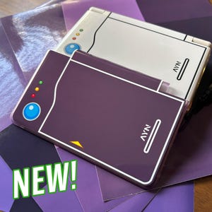 May include: A dark purple handheld gaming console skin featuring a blue circular button, three small status lights, and the text AYN printed in white. A second white console skin with identical design elements is positioned partially underneath the purple one.
