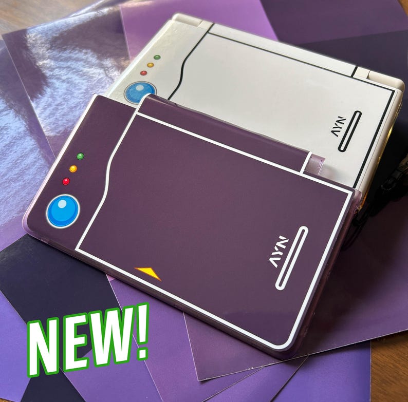 May include: A dark purple handheld gaming console skin featuring a blue circular button, three small status lights, and the text AYN printed in white. A second white console skin with identical design elements is positioned partially underneath the purple one.