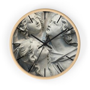 May include: A round wall clock with a light wood frame and black hands. The clock face features a detailed grayscale image of three sculpted stone faces with flowing hair, positioned close together and looking upward in a unified composition.
