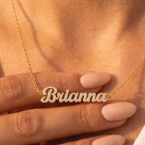 May include: A gold-toned necklace featuring a script-style nameplate pendant that reads Brianna. The nameplate is encrusted with small, clear, sparkling stones and is suspended from a delicate, thin gold-toned chain with a classic link design.
