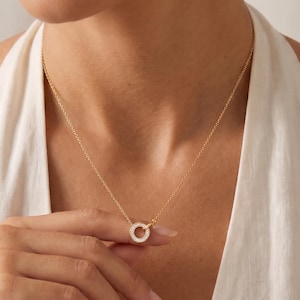 May include: A gold-toned necklace featuring a delicate chain and a circular pendant. The pendant is encrusted with small, clear, sparkling stones along its entire circumference, creating a continuous shimmering effect around the centre ring.