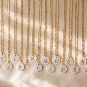 May include: A collection of gold-toned chain necklaces, each featuring a circular white mother-of-pearl pendant engraved with a single uppercase letter. The letters F, G, H, I, J, K, L, M, N, O, P, Q, R, S, T, and U are displayed on individual pendants.