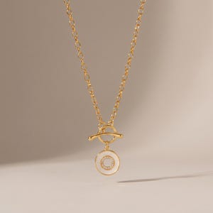 May include: A gold-toned chain necklace featuring a toggle clasp closure. A circular pendant hangs from the toggle, displaying a white enamel border surrounding a smaller inner circle set with tiny clear stones, creating a layered effect.