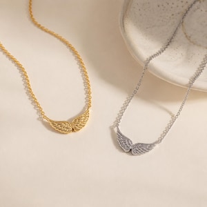 May include: Two necklaces with delicate chains, one in a gold tone and one in a silver tone. Each necklace features a central pendant shaped like a pair of symmetrical angel wings with detailed feather textures, suspended from a thin metal link chain.