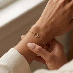 May include: A delicate gold chain bracelet featuring a small, circular charm with a single embedded clear stone at its center. The bracelet is secured with a fine link chain that wraps around the wrist, showcasing a minimalist and refined design.