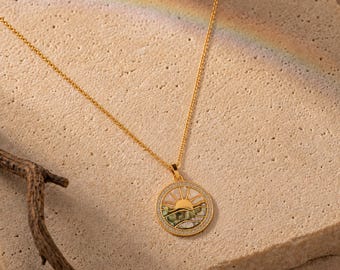 May include: A gold chain necklace featuring a circular pendant with a sun and wave motif. The pendant is inlaid with iridescent shell material and bordered by small clear stones. The design depicts a rising sun over stylized ocean waves.