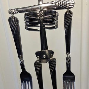 May include: A hanging sculpture shaped like a human figure constructed from stainless steel cutlery. The head is a spoon, the torso is a ribcage made of bent fork tines, and the arms and legs are composed of forks connected by metal jump rings.