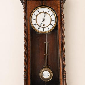 May include: A dark brown wooden wall clock featuring a circular white face with black Roman numerals and ornate hands. The clock has twisted side columns and a glass-fronted lower section displaying a brass pendulum with the letters R and A.