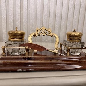 May include: A desk set featuring a polished dark wood base with gold-toned metal accents, two clear glass inkwells with brass lids, a central decorative gold-toned handle, and a single brown quill pen resting across the front of the base.
