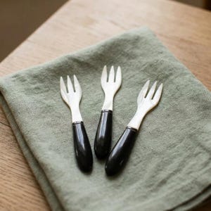 以下が含まれることがあります： 3本の白い三つ又のヘッドと、滑らかで光沢のある黒いハンドルを備えた小さなフォーク3本。フォークは一列に並べられており、明るい色の歯と暗い単色のハンドルのコントラストが際立っており、小さな食べ物を取り分けるのに適しています。