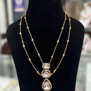 May include: A gold-toned necklace and earring set featuring clear, faceted stones. The necklace has a double-layered chain with small gold beads and a central pendant consisting of three stacked, stone-encrusted frames. Matching teardrop earrings.