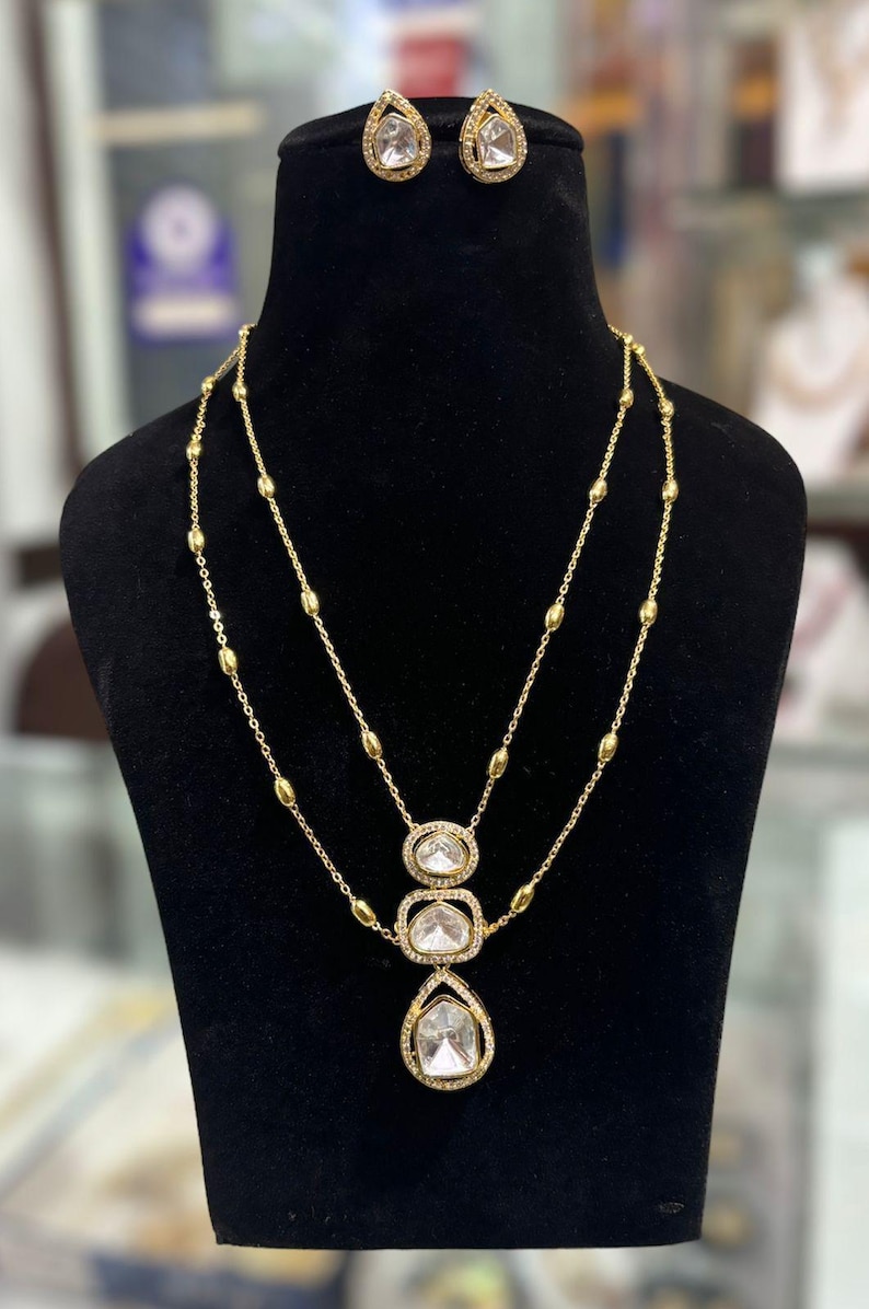 May include: A gold-toned necklace and earring set featuring clear, faceted stones. The necklace has a double-layered chain with small gold beads and a central pendant consisting of three stacked, stone-encrusted frames. Matching teardrop earrings.