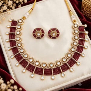 May include: A necklace and earring set featuring multiple strands of small, deep red beads. The necklace includes gold-toned circular accents with white stones and dangling teardrop-shaped pendants. The matching earrings are circular with red beads.