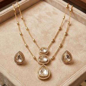 May include: A gold-toned necklace and earring set featuring three teardrop-shaped pendants on a delicate chain with small oval beads. The jewelry pieces are accented with clear, faceted stones surrounded by tiny white crystals in a pav&eacute; setting.
