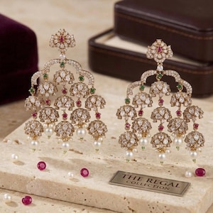 May include: A pair of gold-toned chandelier earrings featuring multiple bell-shaped drops, clear stones, small red and green accents, and dangling white pearls. A metal plaque reads THE REGAL COLLECTION, accompanied by loose red stones and white pearls.