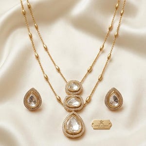 May include: A gold-toned necklace and earring set featuring clear, pear-shaped stones surrounded by small white crystals. The necklace has a double-layered chain with oval accents and a pendant with three stacked stones. A small gold tag reads JK JAIPUR SUNDAN HERITAGE.